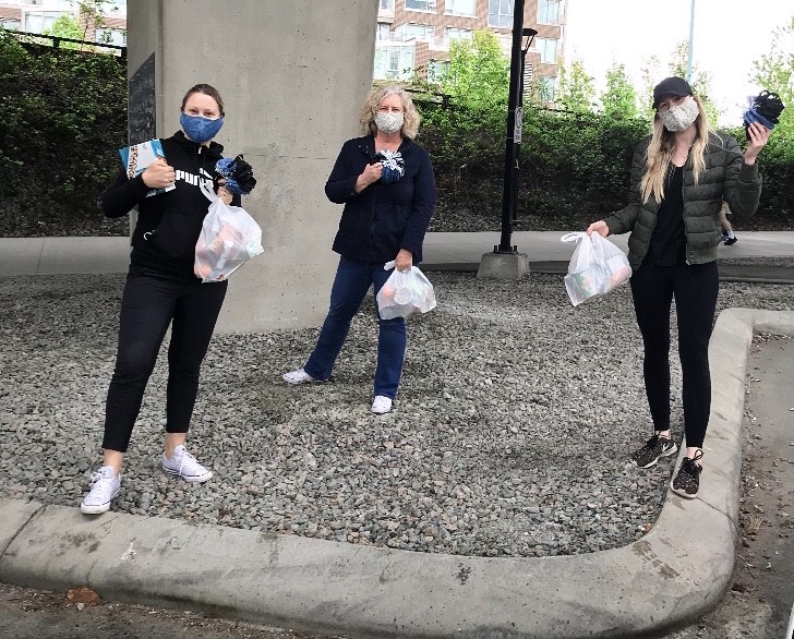 Three masked women standing in a gravel area holding bags of groceries